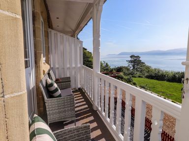 A balcony with wicker chairs and cushions overlooking a garden and a body of water at Menai View near Menai Bridge