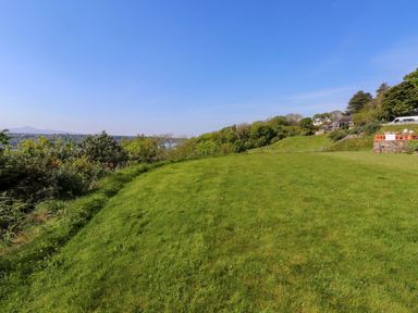 A grassy open field with trees and houses in the background under a clear sky at Menai View near Menai Bridge