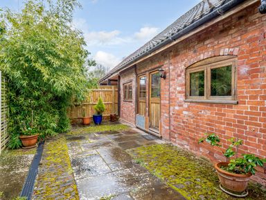 An outdoor patio area with stone paving and moss next to a brick building with wooden doors and windows at The Bothy in Phocle Green near Ross-on-Wye