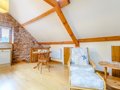 An attic room with wooden beams a small dining table and chair a cushioned armchair with footstool and a side table at The Bothy in Phocle Green near Ross-on-Wye