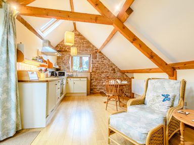 An attic kitchen with wooden beams a dining table two chairs a rattan armchair with ottoman and a stone wall at The Bothy Phocle Green near Ross-on-Wye