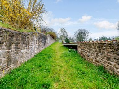 A grassy path between two stone walls with trees and a wooden gate at The Bothy in Phocle Green near Ross-on-Wye