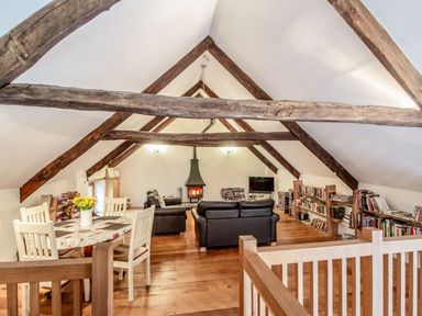 An attic living room with exposed wooden beams a dining table with chairs sofas a wood stove and bookshelves at The Milking Parlour in Highampton