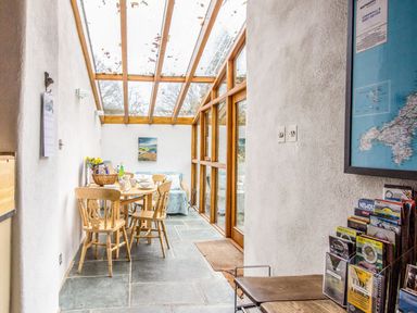 A dining area with a wooden table and chairs beneath a glass roof and wooden framed windows at The Milking Parlour in Highampton