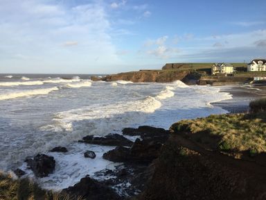 A coastal scene with waves crashing on rocks and a cliff with houses in the background at The Milking Parlour in Highampton