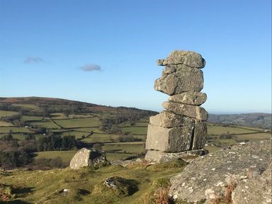 A rock formation balancing on a grassy hill with a landscape of fields and hills under a clear sky at The Milking Parlour in Highampton