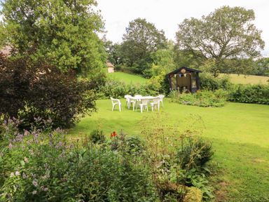 A garden with plastic chairs and a table near a small wooden summerhouse surrounded by plants and trees at The Milking Parlour in Highampton