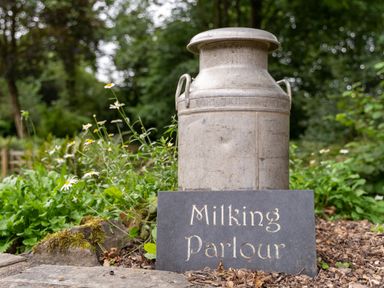 An old milk churn and a sign reading Milking Parlour outdoors with plants and flowers at The Milking Parlour in Highampton