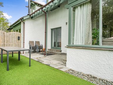 An outdoor area with a table, stacked chairs, a wooden gate, and a green grassy surface at Beech - Woodland Cottages in Bowness-on-Windermere