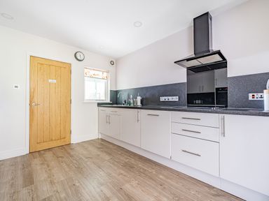 A kitchen with white cabinets, a black countertop, a stove with a black hood, a wooden door, and a window with a patterned blind at Beech - Woodland Cottages in Bowness-on-Windermere