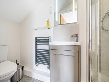 A bathroom with a toilet a sink with cabinet a glass shelf with soap a towel radiator and a shower at Beech - Woodland Cottages in Bowness-on-Windermere