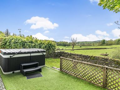 An outdoor hot tub with steps next to a stone wall and wooden lattice fence overlooking green fields at Beech - Woodland Cottages in Bowness-on-Windermere