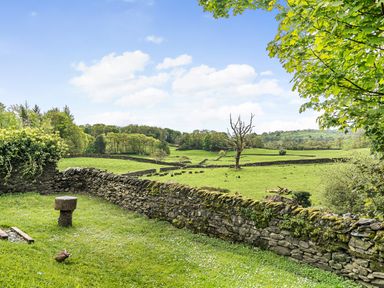 A grassy garden area with stone walls and a tree stump seat overlooking green fields with stone fences and trees at Beech - Woodland Cottages in Bowness-on-Windermere