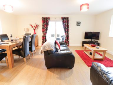 A living room with black leather sofas a wooden dining table with black chairs a coffee table a television and red and black curtains at Arnant in Barmouth