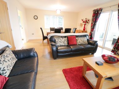 A living room with black leather sofas, a wooden coffee table, a red rug, dining table with black chairs, and floral curtains at Arnant in Barmouth