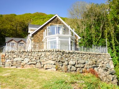 A stone house with glass balcony and stone retaining wall surrounded by trees at Arnant in Barmouth