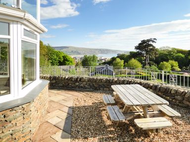 An outdoor patio with a wooden picnic table and benches overlooking a stone wall and greenery at Arnant in Barmouth