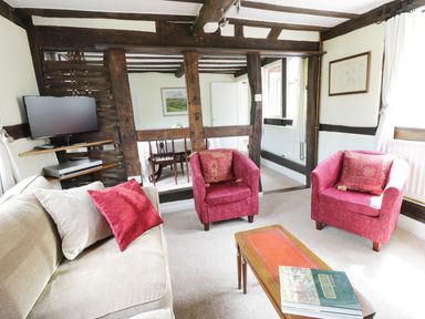A living room with a beige sofa and two red armchairs a wooden coffee table with books a wall-mounted TV and wooden beams at Causeway Cottage in Pencombe