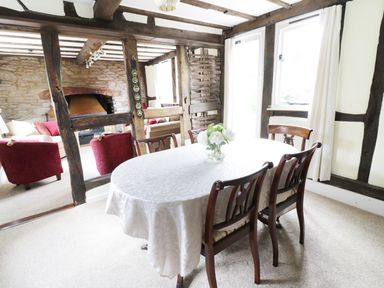 A dining area with a table covered by a white cloth and four wooden chairs next to a room with red armchairs and a stone fireplace at Causeway Cottage in Pencombe