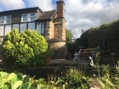 A garden with a stone chimney house bushes a bench a statue and a pond at Causeway Cottage in Pencombe