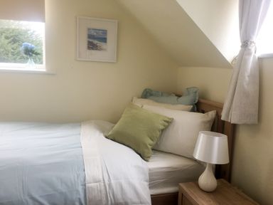 A small bedroom with a single bed next to a wooden bedside table with a white lamp and a window with a curtain and flowers in a vase at Causeway Cottage in Pencombe