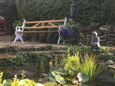 A garden area with a bench, a hanging flower pot, a stone statue of a girl, and a small pond with aquatic plants at Causeway Cottage in Pencombe