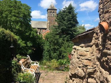 A garden bench on a stone path near a stone building with trees and a church tower in the background at Causeway Cottage in Pencombe