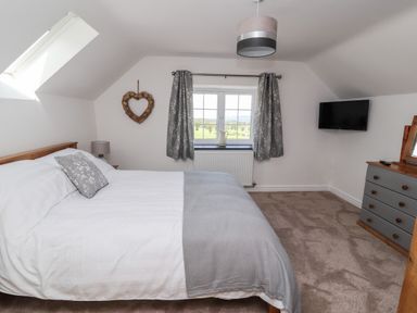 A bedroom with a bed a bedside lamp a heart-shaped wall decoration a window with curtains a wall-mounted TV and a dresser with a mirror at Delfryn Llannefydd near Denbigh