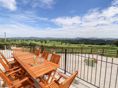 An outdoor wooden table with chairs and a bottle with glasses on a patio overlooking green fields and hills at Delfryn in Llannefydd near Denbigh