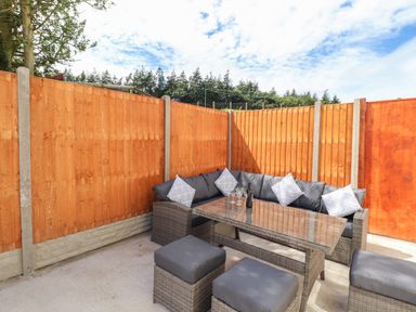 An outdoor seating area with a cushioned wicker sofa table and stools surrounded by wooden fencing at Delfryn in Llannefydd near Denbigh