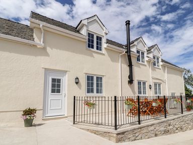 The exterior of a cream-colored house with a patio, wooden furniture, and hanging flower pots at Delfryn in Llannefydd near Denbigh