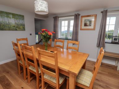 A dining room with a wooden table and eight chairs with cushions a vase of flowers on the table paintings on the walls and windows with curtains at Delfryn in Llannefydd near Denbigh
