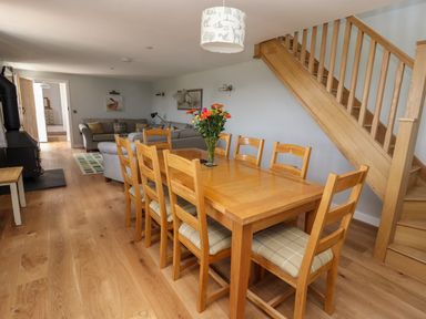 A dining area with a wooden table and eight chairs next to a wooden staircase and a living room with grey sofas at Delfryn in Llannefydd near Denbigh