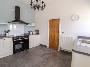 A kitchen with a black stove, white cabinets, wooden door, wall clock, and grey countertops at Delfryn in Llannefydd near Denbigh