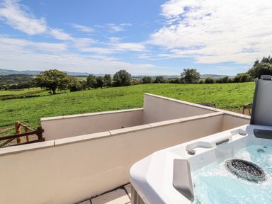 An outdoor hot tub on a tiled patio overlooking green fields and trees at Delfryn in Llannefydd near Denbigh