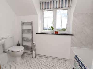 A bathroom with a toilet, a towel rack, a window with plants on the sill, and a bathtub with a striped towel at Delfryn in Llannefydd near Denbigh