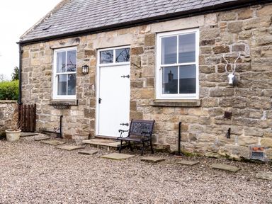 The exterior of a stone building with a white door and two windows a bench and a decorative deer head on the wall at The Old School Room in Longhorsley near Morpeth and Alnwick