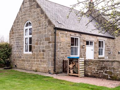 A stone house with a slate roof and a wooden log store with firewood in a garden at The Old School Room in Longhorsley near Morpeth and Alnwick