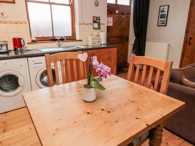 A kitchen with a wooden table and chairs a pink flower in a pot on the table a washing machine and dryer under the counter a window and wooden door in the background at The Old School Room in Longhorsley near Morpeth and Alnwick