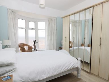 A bedroom with a white bed near a bay window with wicker chair and telescope and a mirrored wardrobe at Number Fifteen in Withernsea