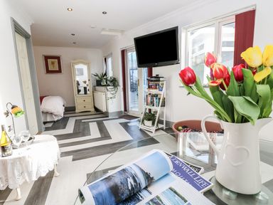 A living area with a glass table holding magazines and a jug of tulips a small side table with wine glasses and a bottle a wall-mounted TV a bookshelf with books and plants a doorway to a bedroom at Chapel Green Studio Polgooth near St Austell