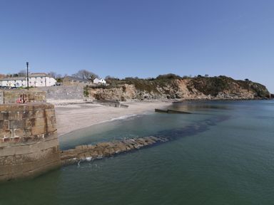 A view of a stone pier and beach with houses and a rocky hill in the background at Chapel Green Studio in Polgooth near St Austell