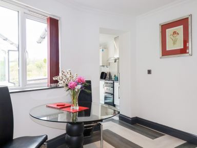 A dining area with a glass round table and two black chairs near a window showing a kitchen in the background at Chapel Green Studio in Polgooth near St Austell