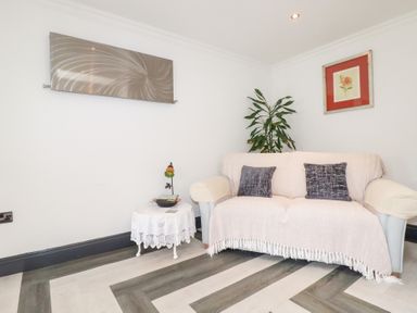 A small living room with a sofa covered with a light throw and two dark cushions a side table with a lace cloth a plant and two wall artworks at Chapel Green Studio in Polgooth near St Austell