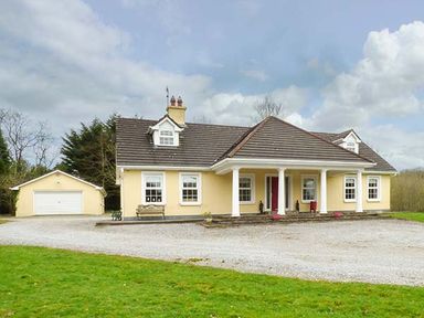 A yellow house with a gravel driveway and detached garage surrounded by grass at The Sanctuary in Killavullen near Mallow