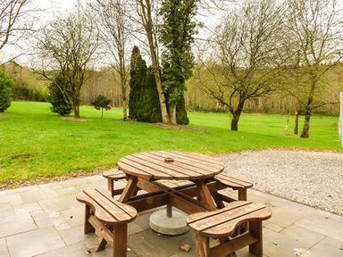 A wooden round picnic table with attached benches on a paved area overlooking a grassy garden with trees at The Sanctuary in Killavullen near Mallow