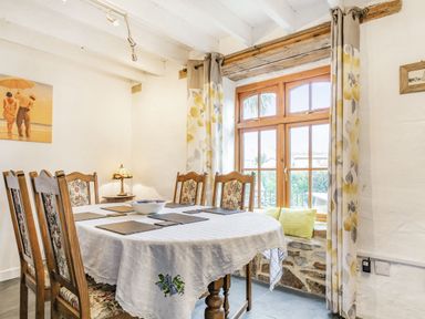 A dining room with a wooden table covered with a white cloth and floral chairs near a window with floral curtains at Victoria Cottage in St Newlyn East near Newquay