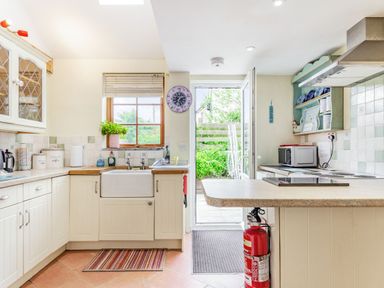 A kitchen with cream cabinets a white sink a microwave and an open door leading outside at Victoria Cottage in St Newlyn East near Newquay