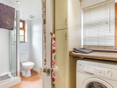 A bathroom with a toilet and shower next to a laundry area with a washing machine and window blinds at Victoria Cottage in St Newlyn East near Newquay