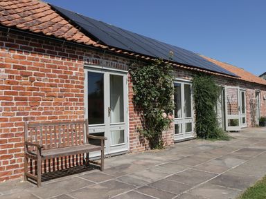A brick building with white framed glass doors and solar panels on the roof with a wooden bench outside at Cow Byre in Malton